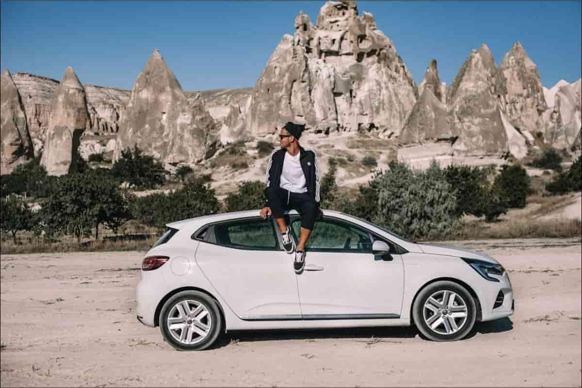 man sitting on his white car in front of hills